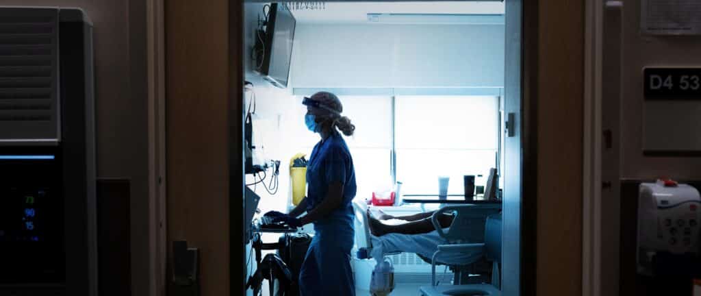 A nurse stands at a computer inside an intensive care unit patient room.